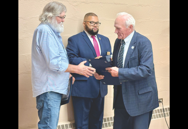 Three men in a ceremony: a bearded man in a navy suit and pink tie hands a folder to an older man in a blue blazer while another man with long gray hair watches.