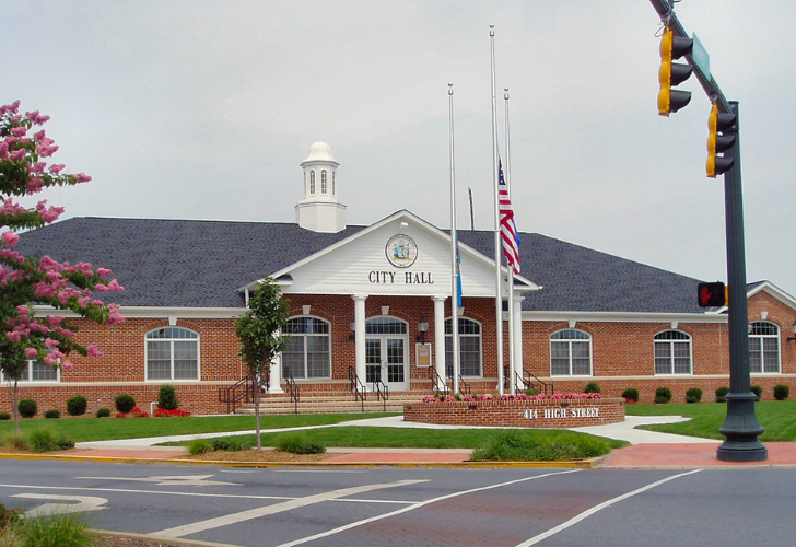 Brick city hall with a white portico entrance and flagpoles in front, across a street from a traffic light.
