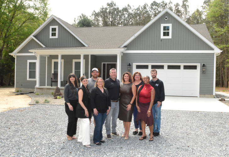 A diverse group of ten adults posing in front of a new gray, two‑car garage house with white trim, a covered front porch, and a gravel driveway.