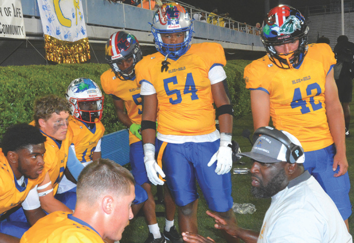 Football players in yellow and blue uniforms gather around a coach on the sideline, listening during a break.