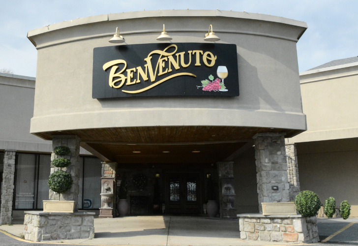 Restaurant entrance with a curved beige façade and a black sign reading 'Benvenuto' above the doorway, flanked by stone columns and greenery.