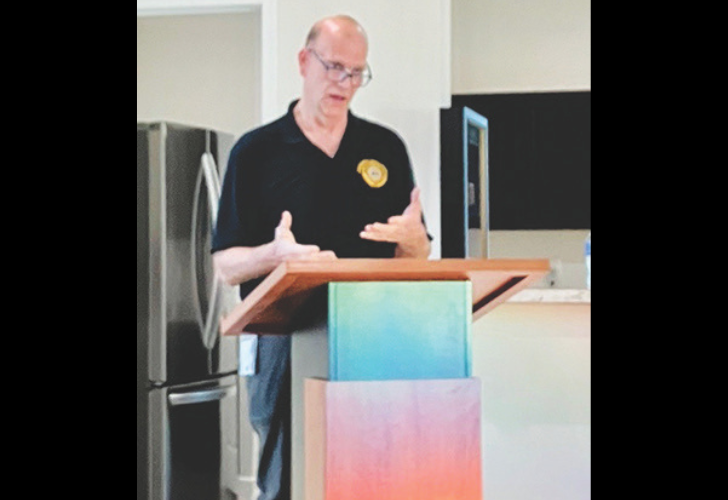 Man giving a talk at a wooden podium in a modern kitchen or office space, gesturing with hands as he speaks.
