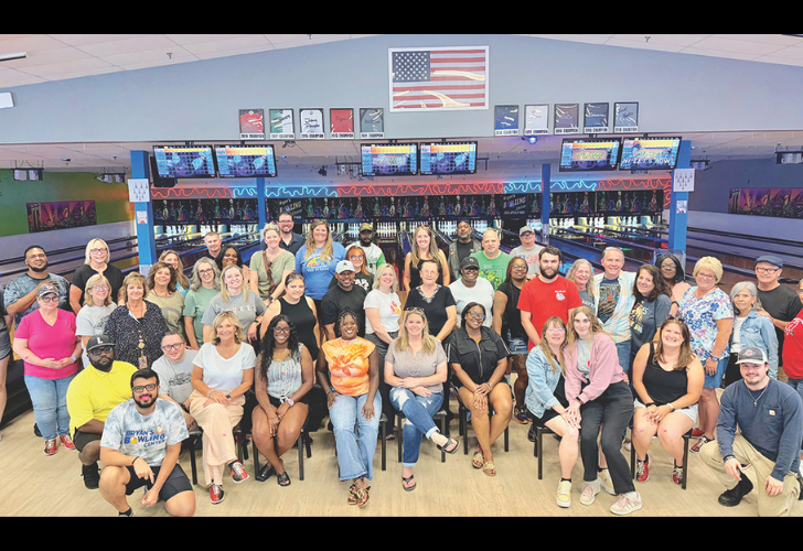 Group of 40+ people posing for a group photo in a bowling alley, with lanes, neon lights, and an American flag on the back wall.
