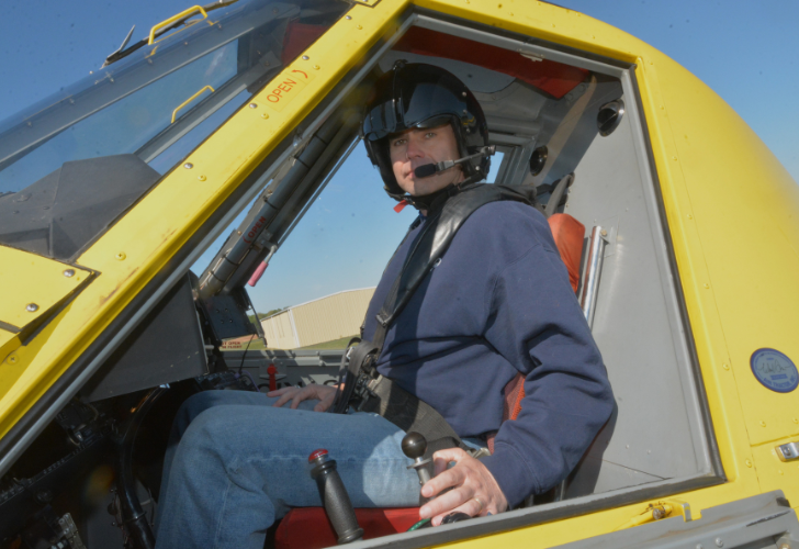 Pilot wearing a helmet and headset sits in the cockpit of a bright yellow aircraft, gripping the control stick with a clear blue sky outside.