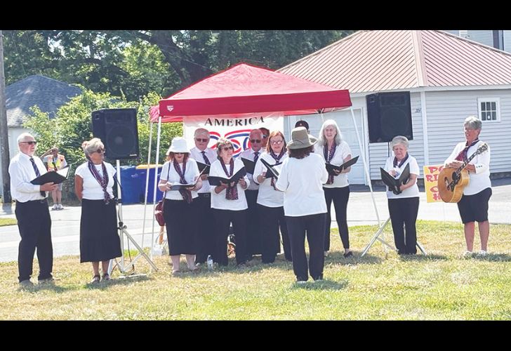 Chorus- page 10 Outdoor choir under a red canopy performing, conductor at front, guitarist on the right, singers in white tops and scarves with a banner reading AMERICA behind them.