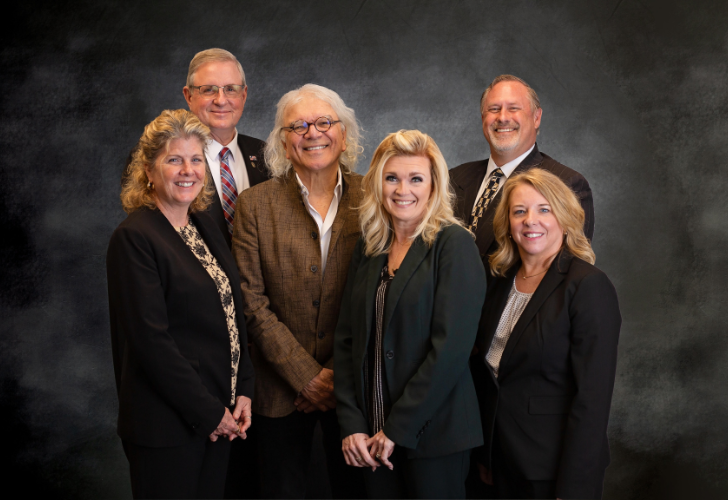 Six professionals in business attire posing for a formal team portrait against a dark backdrop.