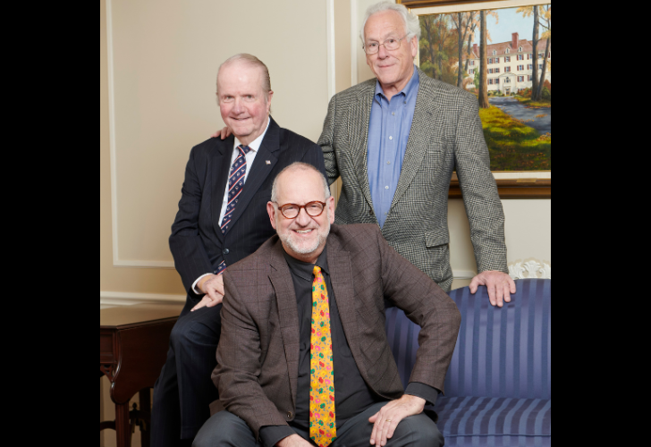Three older men posing for a photo in a living room; the man in front wears a brown blazer and yellow patterned tie.",