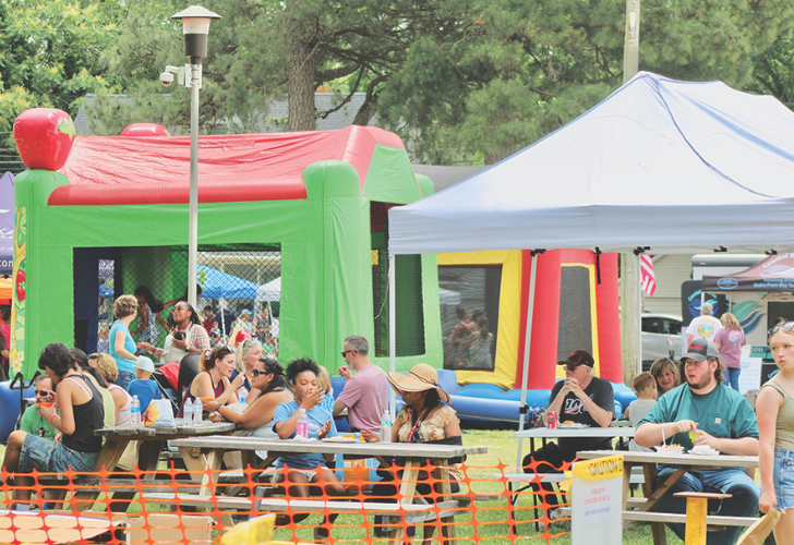 Families and friends sit at picnic tables under a white canopy at an outdoor festival, with colorful inflatable bounce houses in the background.