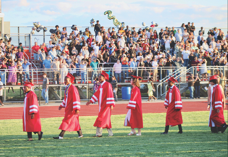 Line of graduates in red gowns and caps walking on a football field as spectators watch from the bleachers at a parade-like ceremony.