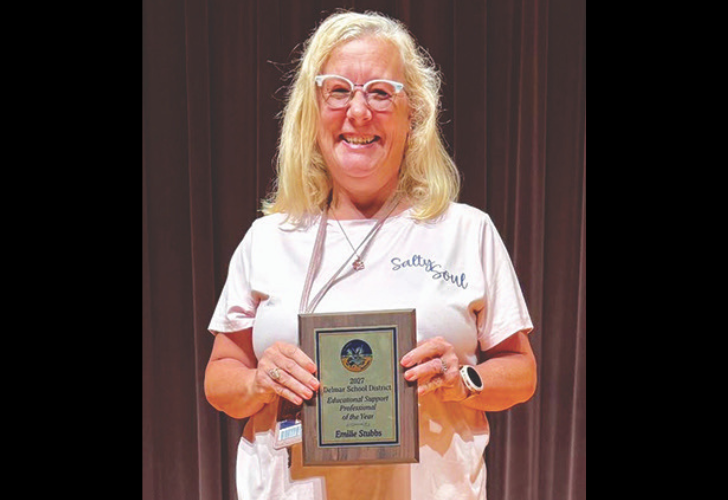 Smiling woman with glasses holding a framed award plaque on a stage with a dark curtain behind her.