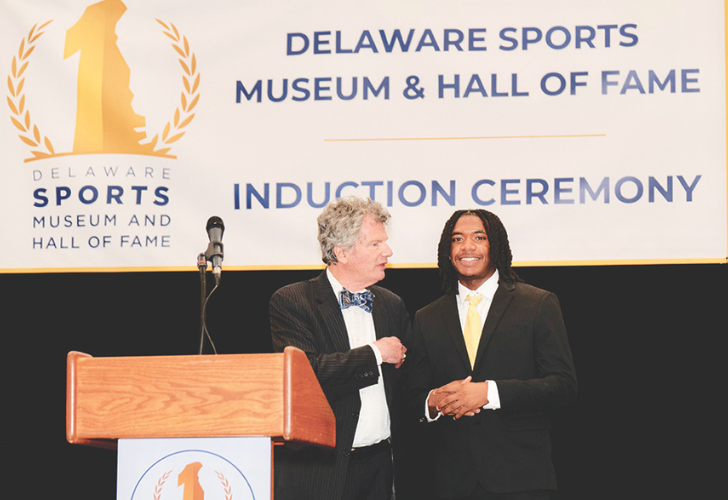 Two men in suits shake hands on a stage at the Delaware Sports Museum induction ceremony.