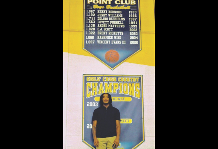 Yellow gym banner with basketball honors and a shield listing players’ names, above a 'Girls' Cross Country Champions 2003' banner where a man stands in front.