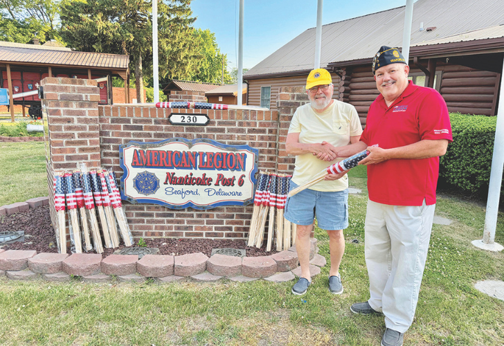 Flag presentations- page 30 Two men stand by a brick wall with an American Legion Post 6 sign, holding a rolled American flag with patriotic staffs nearby.
