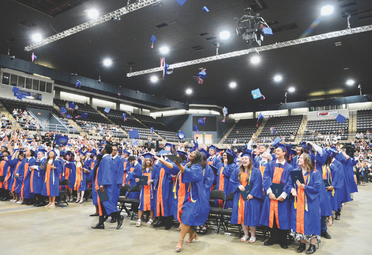 Group of graduates in blue gowns in an arena, tossing mortarboards as cheering spectators look on.