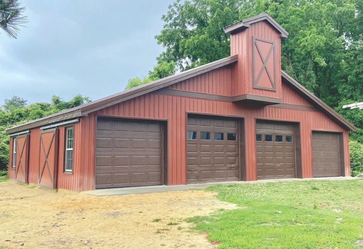 Red barn-style garage with four brown doors, a small cupola with an X on top, and trees in the background.