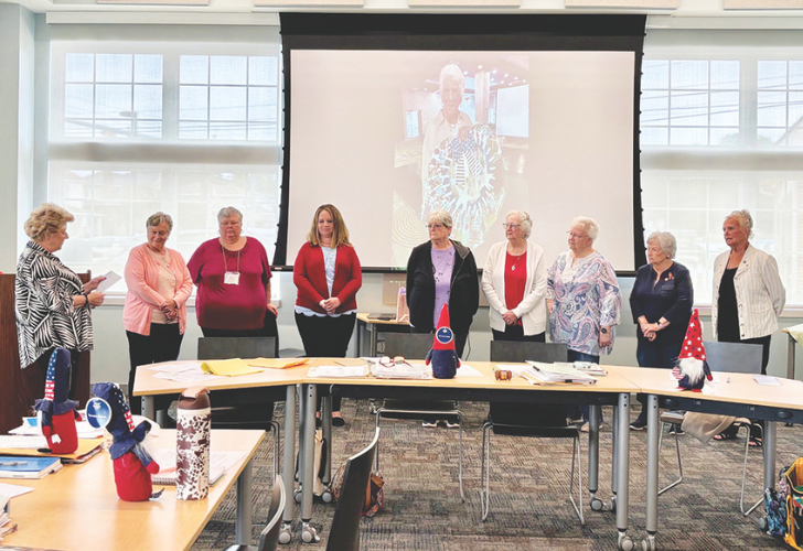 Group of senior women standing along a long conference table in a bright room, projector screen with a person’s image behind them.