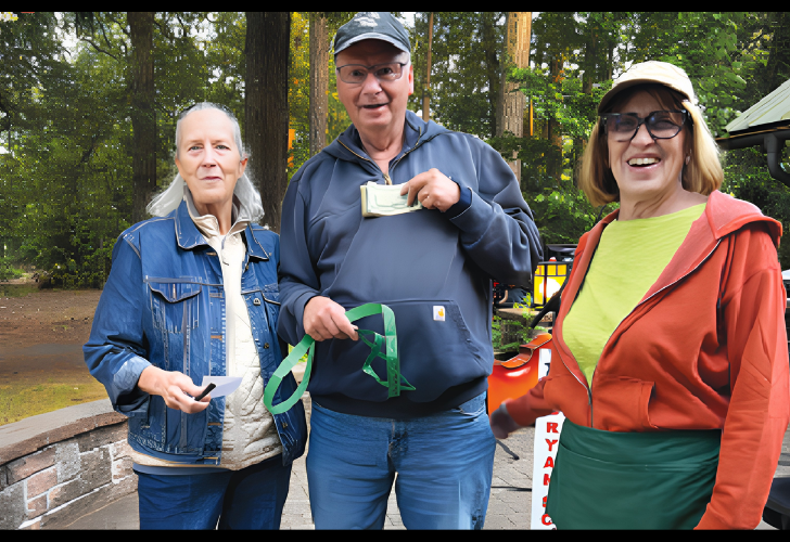 Three smiling adults outdoors in a park; the man in the center holds cash and a green ribbon, while the woman on the left holds a card and the woman on the right wears an orange jacket and cap.
