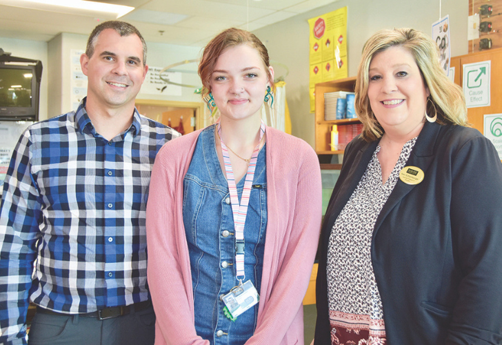 Three smiling coworkers pose together in a bright workplace; the woman in the center wears a denim outfit and ID badge on a lanyard.