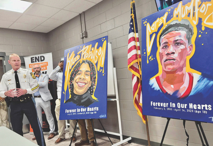 Memorial display: two large portraits on easels—one of a smiling woman and one of a basketball player—titled 'Forever In Our Hearts' with an American flag nearby, a police officer and others in the background.