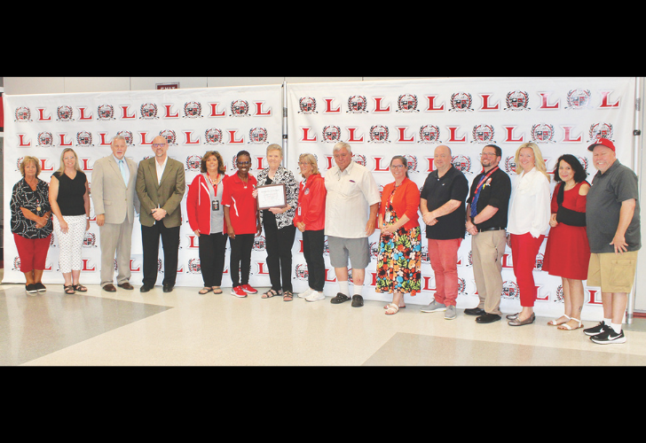 Group of about 16 people standing in a row, one person holding a framed certificate, in front of a backdrop with repeated L logos.