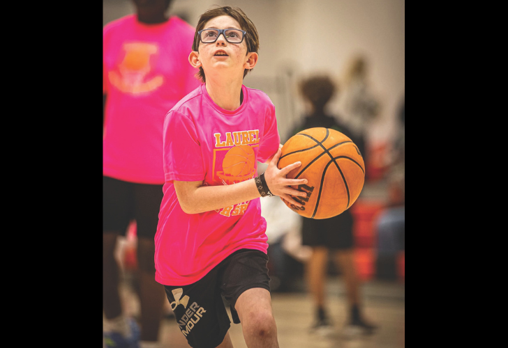 Child in a pink t‑shirt dribbling a basketball indoors, wearing glasses and focused on the play.