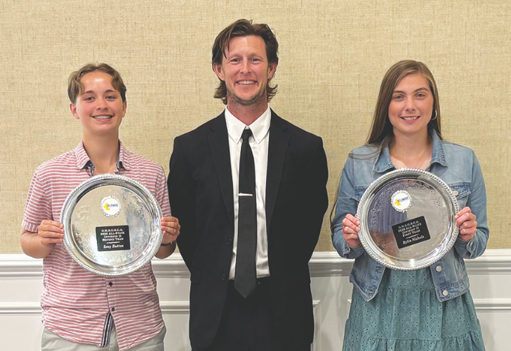 Three people stand in a row; two teens hold silver plate awards while a man in a black suit smiles between them in a formal setting.