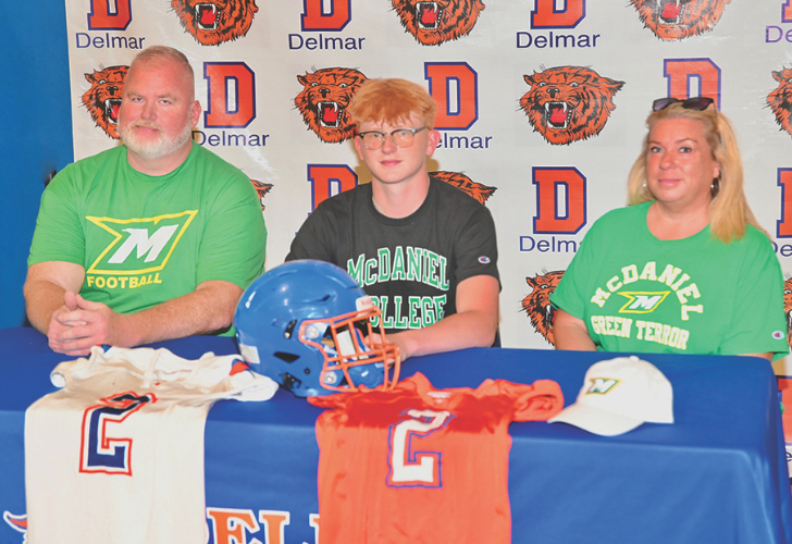Three people sit at a signing table in front of a Delmar backdrop, football gear and jerseys displayed on the table, including a McDaniel College shirt wearer in the middle and a green-teal shirt on the right.