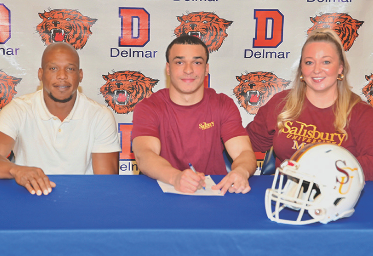 Three people sit at a blue table during a signing ceremony; the student in the middle signs a document while two adults flank him, with a backdrop of Delmar logos and tiger mascots and a Salisbury football helmet on the table.