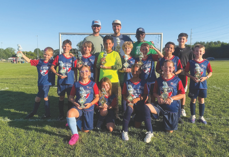 Youth soccer team posing on a sunny field with trophies, flanked by three coaches behind them.