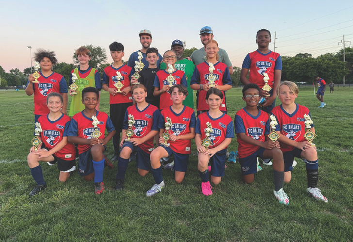 Youth soccer team posing on a grassy field after a tournament, holding trophies; players in red/blue jerseys with FC Dallas logos and coaches behind them.