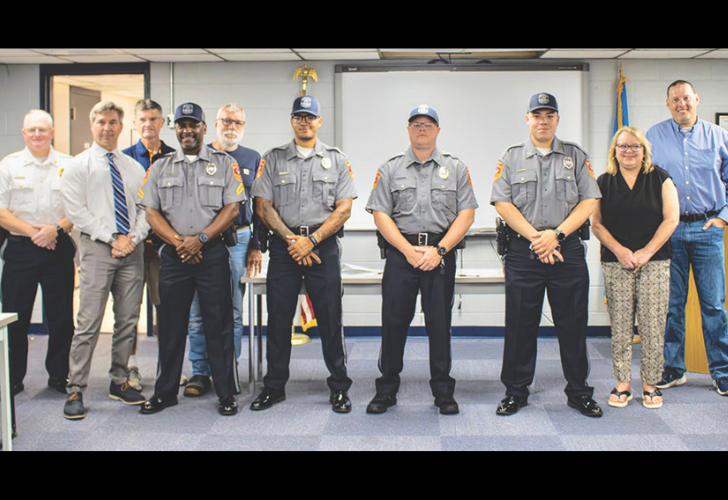 Line of uniformed officers and civilians posing for a formal group photo in a room with a projector screen and flag in the background.