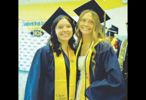 Two smiling graduates in navy gowns with gold stoles and mortarboards posing together at a Class of 2026 graduation.
