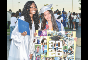 Two graduates in silver gowns and caps pose outdoors, one holding a diploma and the other displaying a 2026 collage board with photos.