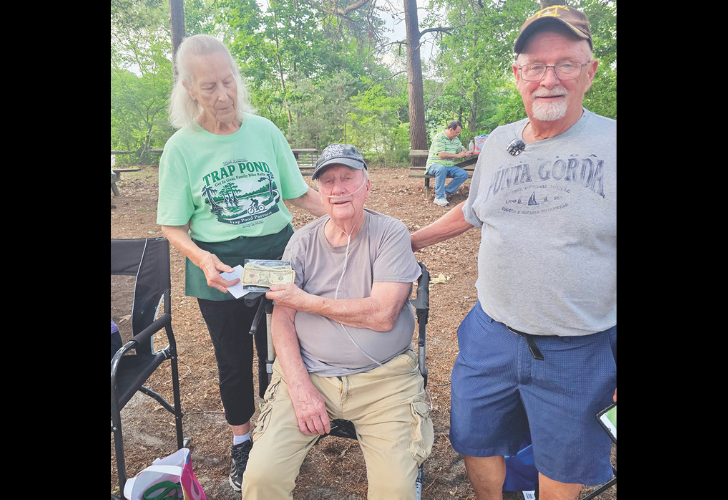 Summer Concert- page 18 Three seniors outdoors in a park; a man in a wheelchair accepts cash from a smiling woman while another man stands beside them, all casually dressed.