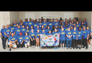 Group of people in matching blue shirts posing for a group photo indoors, holding a colorful USA Games banner.