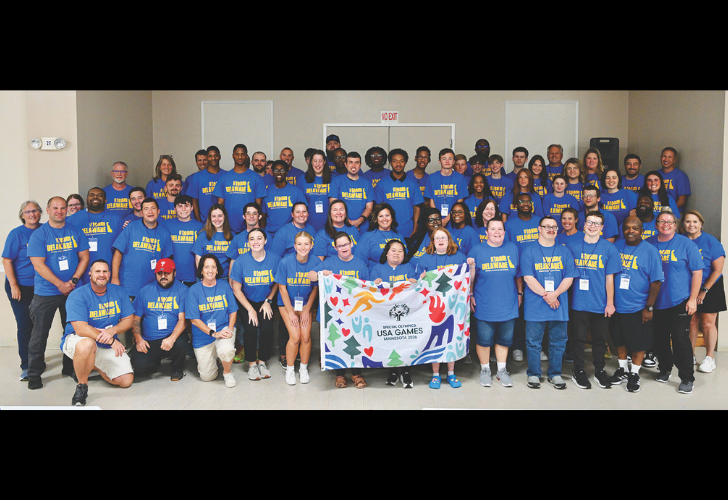 Group of people in matching blue shirts posing for a group photo indoors, holding a colorful USA Games banner.