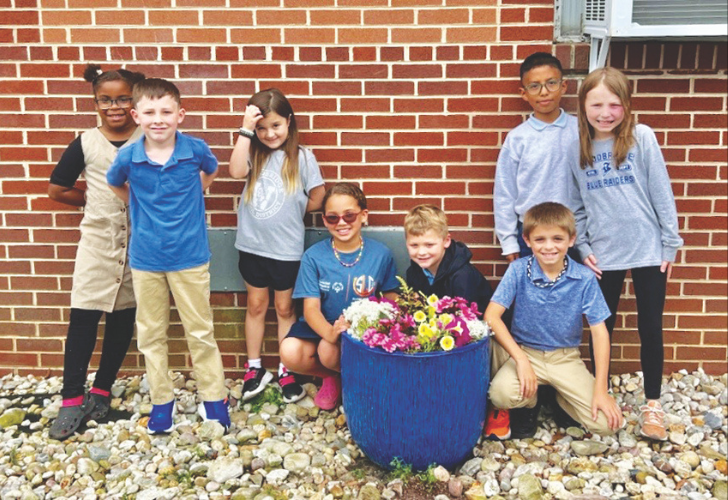 WECEC- page 33 Eight children pose together outdoors beside a brick wall, with a blue pot of colorful flowers in front.