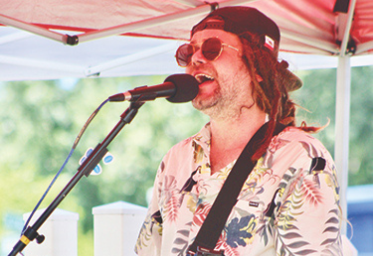 Male singer performing outdoors under a red canopy, wearing a floral shirt, sunglasses, and a backward cap.