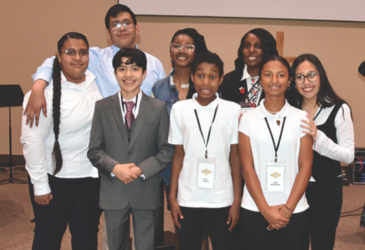 Group of eight students posing together at a school event; some wear lanyards and one boy is in a suit, all smiling.