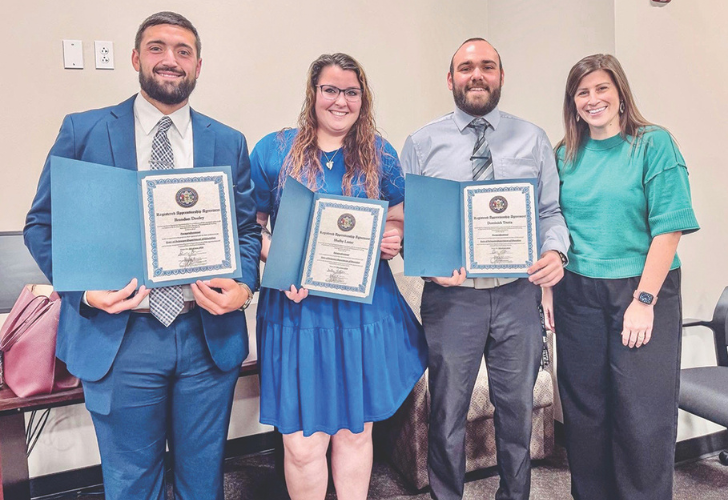 Four adults stand indoors, each holding a blue certificate folder and smiling for the photo.