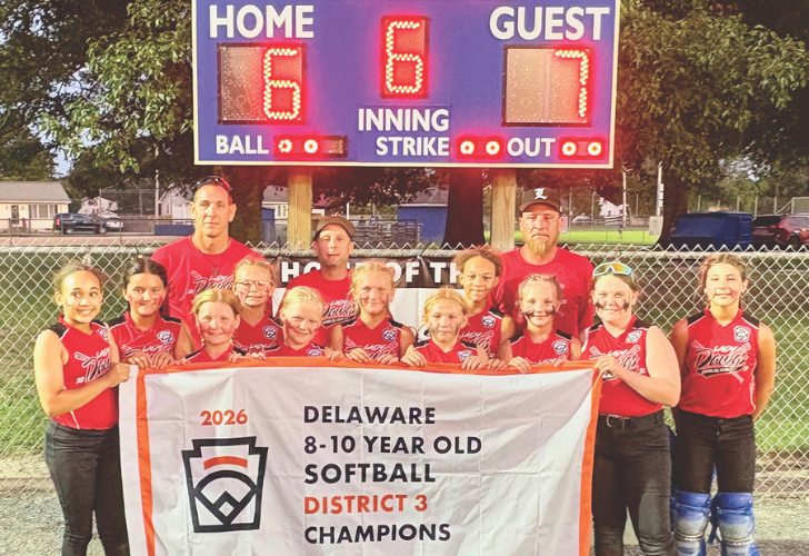 Group of smiling girls in red uniforms pose with a Delaware 8–10 year‑old softball banner announcing District 3 Champions (2026) with a scoreboard in the background.