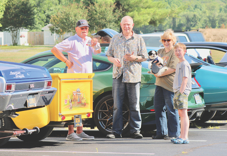 Group of adults and a child posing beside colorful cars in a parking lot, one person holding a small dog in a woman’s arms.