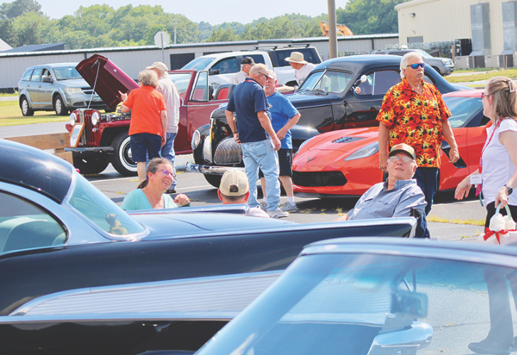 People gather at a car show in a parking lot, admiring various vintage cars and sports cars.
