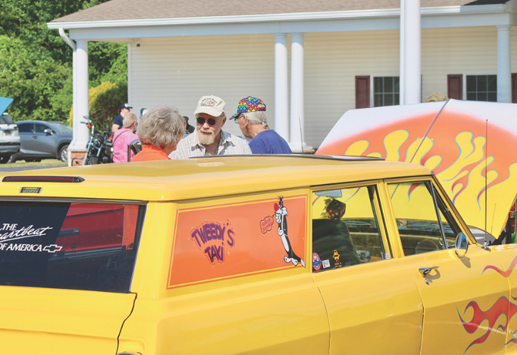 Yellow vintage taxi van with Tweedy's Taxi graphic and flame designs, as people chat at a car show by a light-colored building.