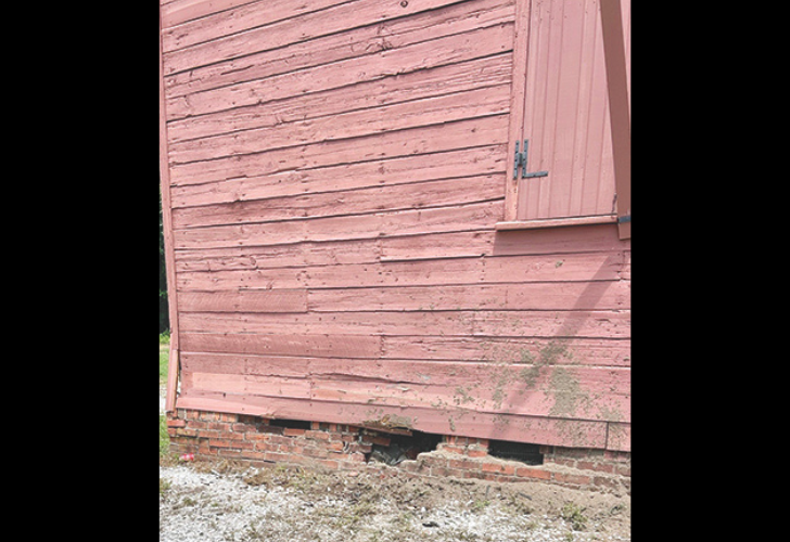 Pink wooden wall of a barn with a cracked brick foundation and gaps at the base, showing weathered siding and moss.