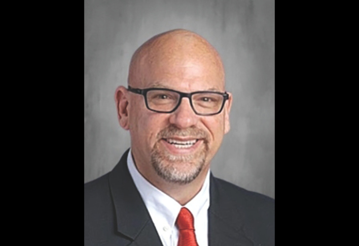 Smiling bald man in a black suit and red tie, wearing glasses, professional headshot against a gray background.