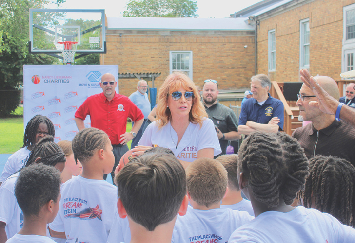 Dream Court Woman with orange hair and sunglasses speaks to a group of children at a charity event outdoors, banner and basketball hoop in view.