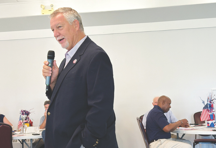 Older man in a dark blazer speaks into a handheld microphone at an indoor event, with seated attendees in the background and small American flags on the tables.
