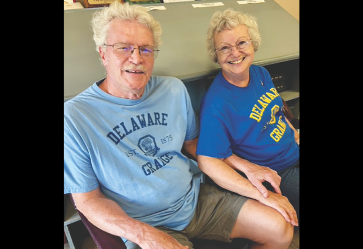 Two smiling seniors sit together, a man in a light blue Delaware Grange shirt and a woman in a blue Delaware Grange shirt, indoors at a counter.
