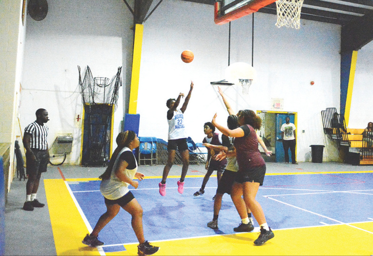 Jump shot- Seaford 30- 7_9 pix Women playing basketball in an indoor gym; a player in white jumps to shoot while defenders reach up near the basket, with a referee watching on the left.